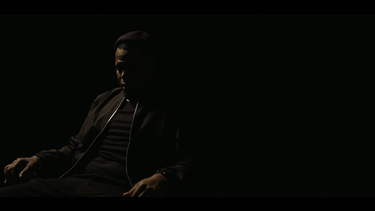 A seated figure in a dark jacket, lit from one side in an otherwise black room