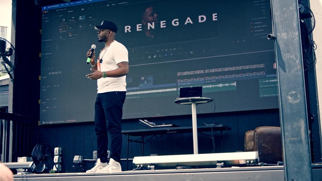 Leon Cuttem stands mid-sentence with a handheld microphone on an outdoor stage, backed by a wall-size screen showing a DaVinci Resolve timeline with the word RENEGADE laid across the viewer.
