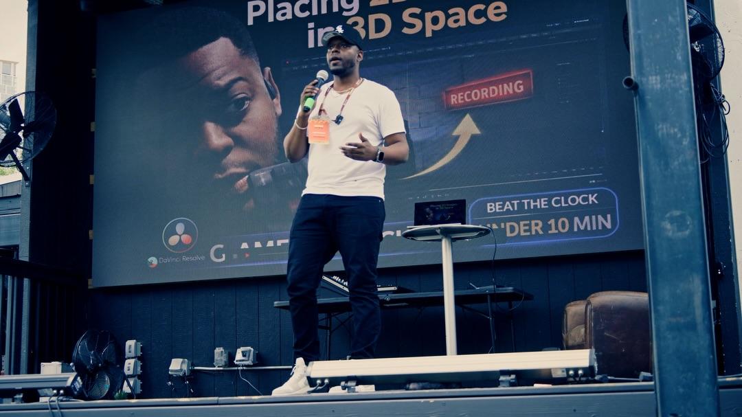 Leon Cuttem gestures while speaking into a handheld microphone on an outdoor stage, the screen behind him showing a presentation slide about placing footage in 3D space, marked with a red RECORDING tag and a “Beat the clock — under 10 min” badge.